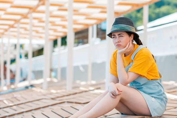 Emotion of sadness and discontent. Portrait of young woman in a straw hat leaned on her hand in discontent, sitting in the shade on the sun beds.