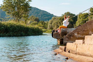 Yaz ve tatiller. Mutlu, kilolu bir kadın, iskelede otururken poz verir. Arka planda, ağaçlar ve deniz. Boşluğu kopyala.