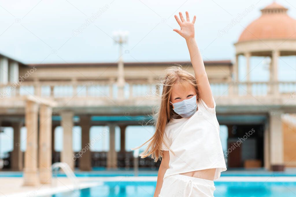 A cute kid in a medical protective mask, joyfully raised her hand up. Outdoor. Concept of protection against the virus and the end of the pandemic.