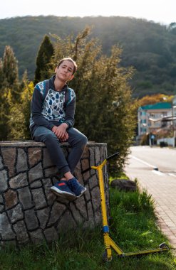 Genç bir çocuk, ayaklarının dibinde bir scooterla taş korkuluğa oturur ve gururla gülümser. Ağaçlar ve arka planda bir sokak. Ergenlik ve gençlik aktivitesi kavramı..