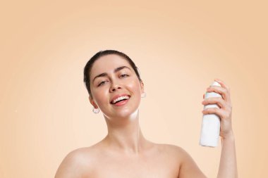 Islak tenli, elinde kozmetik şişesiyle gülümseyen beyaz bir kadın. Uzayı kopyala Bej arka plan. Kozmetoloji, cilt bakımı ve bronzlaşma kavramı.