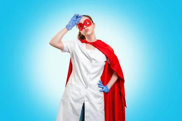 A tired doctor in a mask, medical gloves, and red superhero Cape wipes sweat from her forehead. Blue background. The concept of the Power of a super hero for medicine and stress at work. Copy space.