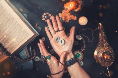 Astroloji ve ezoterizm. Cadı elinde mühür taşlarını tutuyor. Eller yukarı. Siyah bir arka planda fal taşı, bir kitap, değerli tılsımlar, bakır bir lamba ve bir mum yatıyor..