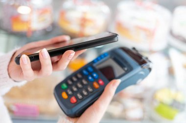 Bir kadının elleri ödeme terminalini tutuyor ve akıllı telefon kullanarak satın almaya para ödüyor. Arka planda pastalar olan bir gösteri. NFC, iş ve bankacılık işlemleri kavramı.