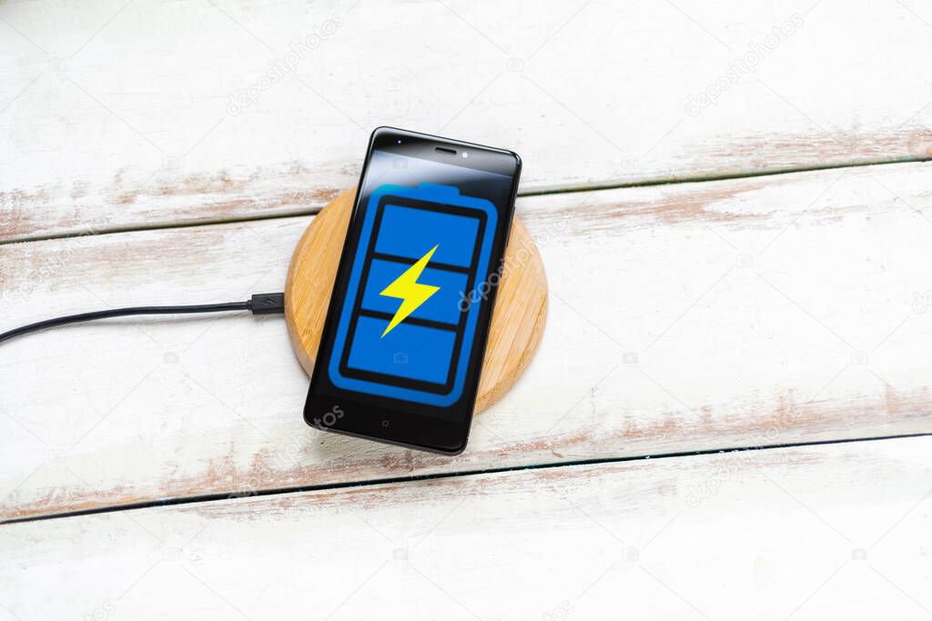 Concept of modern technologies. On the wireless charger is a black smartphone with a blue battery icon. White wooden background with horizontal stripes. Copy space. Flat line.