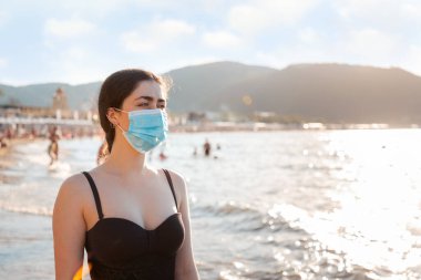 Plajda mayo ve tıbbi maske takmış bir kadının portresi var. Virüs salgını sırasında dinlenme kavramı. Boşluğu kopyala.