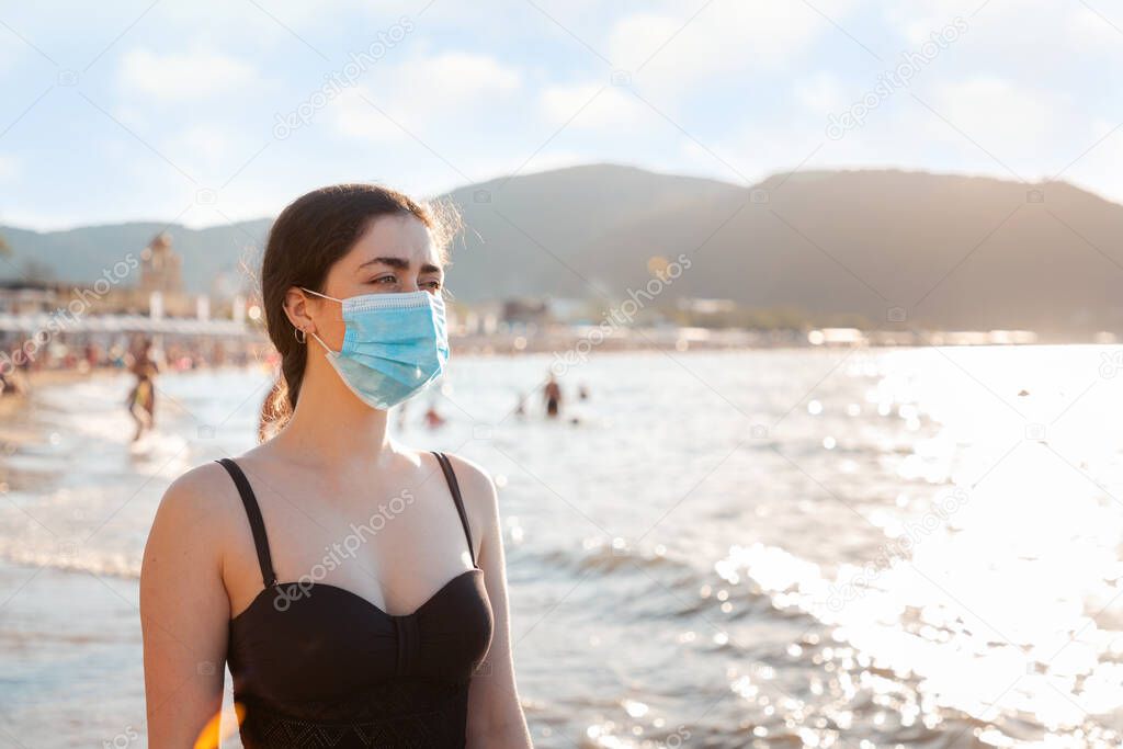Portrait of a woman in a bathing suit and medical mask stands on the beach. Concept of rest during a viral pandemic. Copy space.