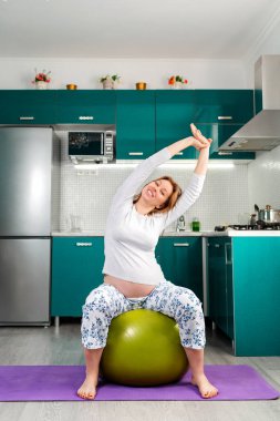 Kendini soyutlama ve evde eğitim. Hamile, beyaz bir kadın bir fitball topunun üzerine oturur ve kollarını uzatır ve sağa doğru eğilir. Mutfak arka planda. Hamile kadınlar için sağlık ve zindelik kavramı.