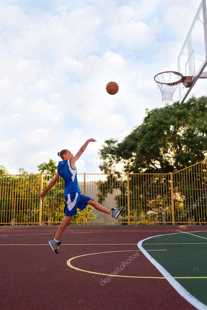 Basketball. A Teenage boy in blue sportswear funny jumps and throws a ball into a basketball Hoop. Sports field in the background. Cloudy sky. Copy space. Concept of sports games.
