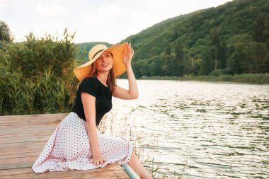 Yaz tatili. Güzel bir genç kadının portresi zarif bir şekilde poz veriyor, deniz kenarında bir iskelede oturuyor ve şapkasını tutuyor. Arkaplanda manzara.