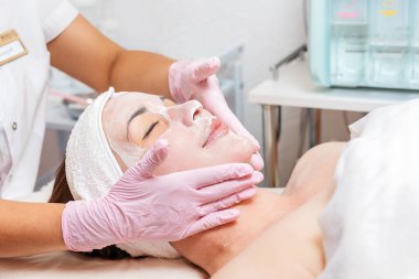 Lastik eldiven takan bir güzellik uzmanı bir kadının yüzüne örülmüş bir maske takar. Memnun bir müşterinin yüzüne yakın çekim. Kozmetoloji ve tedavi kavramı.