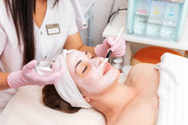 Kozmetik uzmanı, müşterinin yüzüne bir fırça maskesi takar. Kozmetik prosedür. Üst Manzara. Arka planda elektronik bir cihaz var. Kozmetik ve sağlık kavramı.