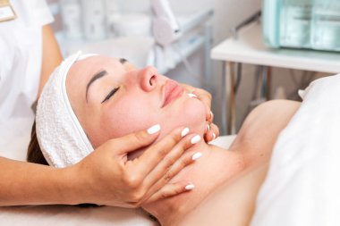 Kozmetik uzmanı bir kadını yüz masajı prosedürü haline getirir. Müşterinin portresi ve doktorun elleri yakın planda. Kozmetik ve güzellik kavramı.