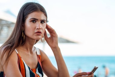 Gergin bir suratı olan bir kadın elinde bir telefon tutuyor. Sahildeki yazlık kafe. İş ve sosyal ağlar kavramı.