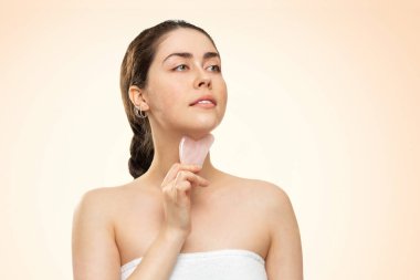 Gua Sha Masajı. Beyaz banyo havlusu giymiş bir kadının portresi boynuna kuvars gua Sha kazıyıcısı ile masaj yapıyor. Bej arka plan. Uzayı kopyala Alternatif tıp ve kozmetoloji kavramı.