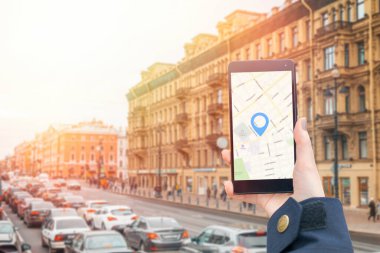 Ceket giymiş bir kadının elinde online harita uygulaması olan bir akıllı telefon var. Arka planda trafik sıkışıklığı olan bir şehir sokağı var. İnternet navigasyon ve modern teknolojiler kavramı.