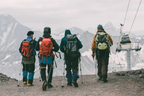 Extreme recreation and mountain tourism. A group of four tourists walking on a mountain trail. In the background there are big snow-covered mountains and a funicular. The view from the back. Close up.