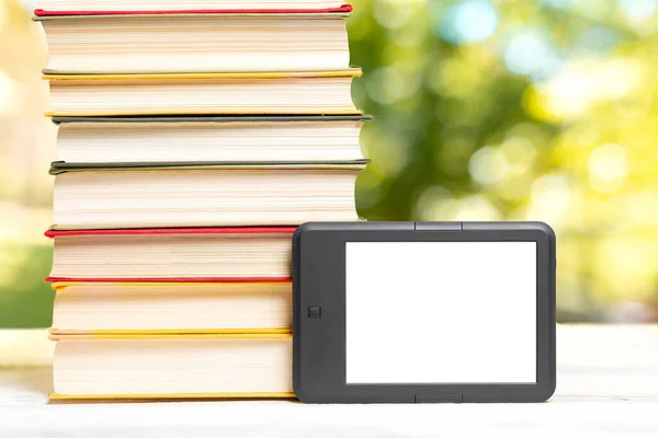 Reading and knowledge. E-book reader and a stack of books. The park is blurred in the background. Mock up. Concept of education and electronic gadgets.