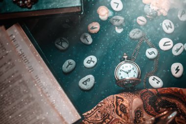Astroloji ve ezoterizm. Karanlık yüzeyde bir zincir üzerinde bir saat, falcı Runik Taşlar, bir kitap ve bir bez bulunur. Yukarıdan manzara. Toz ve ışık.
