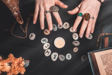 Astroloji ve ezoterizm. Dişi cadı elleri rün çemberini büyülüyor. Siyah arka planda fal baktıran rünler, bir kitap, değerli tılsımlar, bir bakır lamba ve bir mum bulunur. Boşluğu kopyala.