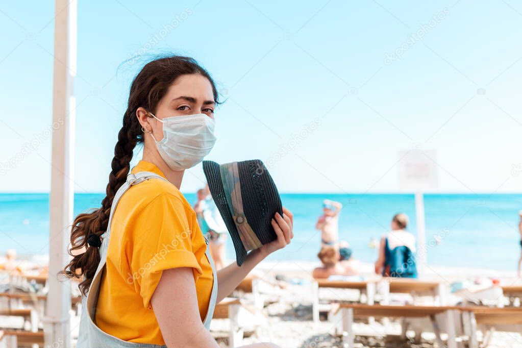 A woman in a medical mask, fanning herself with a hat from the heat. In the background, the beach and the sea. The concept of vacation during a viral pandemic.