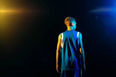 Basketbol mu? Mavi spor kıyafetli genç bir çocuk siyah arka planda duruyor. Çok renkli neon ışıklar. Arkadan arkaya bak. Uzayı kopyala Spor oyunları kavramı.