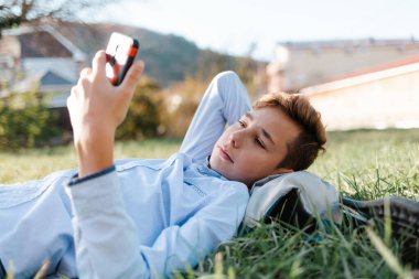 Cep telefonu ve internet. Genç bir çocuk parkta çimlerin üzerinde uzanıyor ve akıllı telefondan video izliyor..