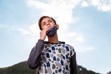 Genç bir çocuk yüzündeki tıbbi maskeyi çıkarıp kıvranıyor. Arka planda gökyüzü. Duygu. Bakterilere ve alerjilere karşı koruma kavramı, koronavirüs salgınına son verir..