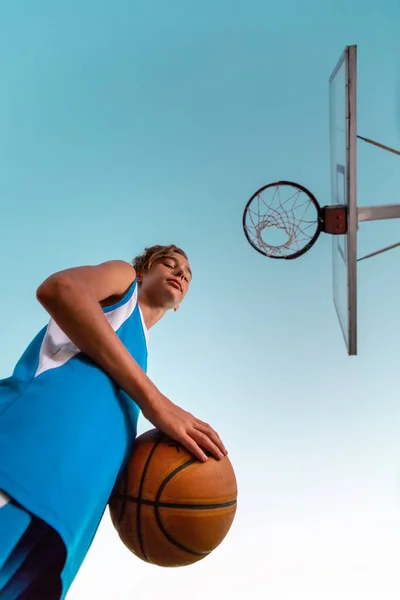 バスケットボールだ 青いスポーツウェアを着た10代の若者がボールを手に持って自信を持って見下ろしています 一番下だ 空を背景に スポーツゲームの概念 — ストック写真