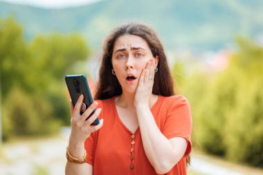 Elinde telefon tutan bir kadının portresi ve korkuyla yüzünü kavraması. Park arka planda. Çevrimiçi dolandırıcılık ve sahte haber kavramı.