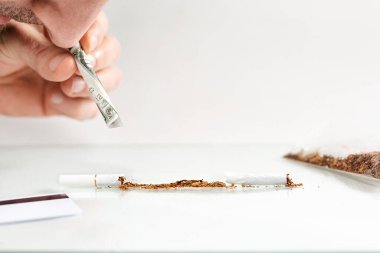 Nikotin bağımlılığı ve sigara karşıtlığı kavramı. Adam sigaradan dökülen tütünü bir dolarlık banknot, sarılmış bir tüp kullanarak soluyor. Beyaz arka plan. Burun ve el yakın..