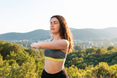 Genç, tatlı bir kadın parkta antrenman yaparken ısınıyor. Yoga, spor ve meditasyon konsepti. Yerleşim yerinin ve dağların arka planında.