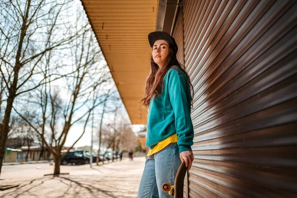 Genç, güzel, hippi bir kadın, eli kaykayın üzerinde poz veriyor. Arka planda kapalı dükkanları olan bir cadde var. Alt manzara. Uzayı kopyala Spor yaşam tarzı ve sokak kültürü kavramı.