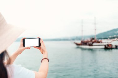 Model yap. Şapkalı kadın telefonu yatay olarak tutuyor. Arka planda, deniz ve gemiler. İzole et.