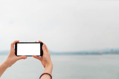 Model yap. Kadın elleri telefonu yatay olarak tutuyor. Arka planda deniz ve gökyüzünde. Uzayı kopyala İzole et.