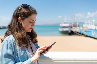 Cep telefonu kullanan güzel bir genç kadın. Arka planda deniz ve gemiler. Boşluğu kopyala. Modern teknoloji ve internet kavramı.
