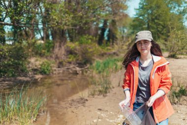 Genç bir kadın gönüllü, çöp torbasında nehrin kenarında çöp ve plastik şişe topluyor. Dünya Günü, ekoloji ve çevre koruma kavramı.