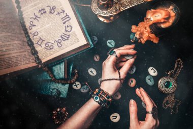 Astroloji ve ezoterizm. Cadı Runik Taşlar hakkında tahmin yürütüyor. Eller yukarı. Siyah arka planda fal baktıran rünler, bir kitap, değerli tılsımlar, bir bakır lamba ve bir mum bulunur. Kitapta Zodiac burcu var..