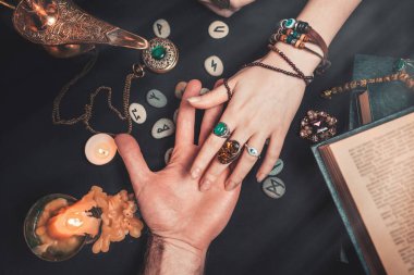 Astroloji ve ezoterizm. Falcı bir adamın avuç içini okur. Siyah arka planda runikler, bir mum, tılsım ve bir kitap var. Yukarıdan görünüşü.