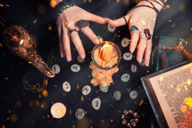 Astroloji ve ezoterizm. Dişi cadı elleri mumun üzerinde hokkabazlık yapar. Siyah arka planda fal baktıran rünler, bir kitap, değerli tılsımlar, bir bakır lamba ve bir mum bulunur. Havadaki ateş kıvılcımları.