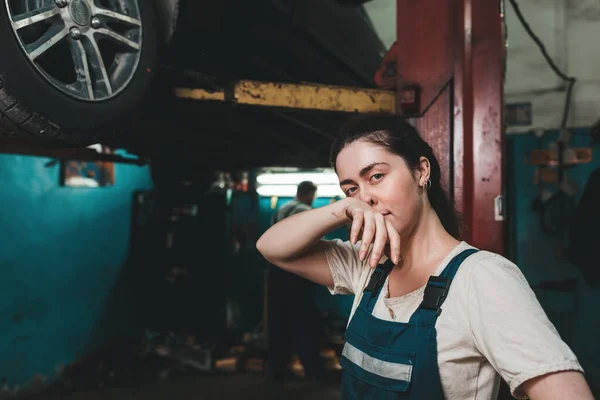 Gender equality. A young brunette in uniform stands near the Elevator with a car and wipes her nose with her hand. In the background is an auto repair shop.