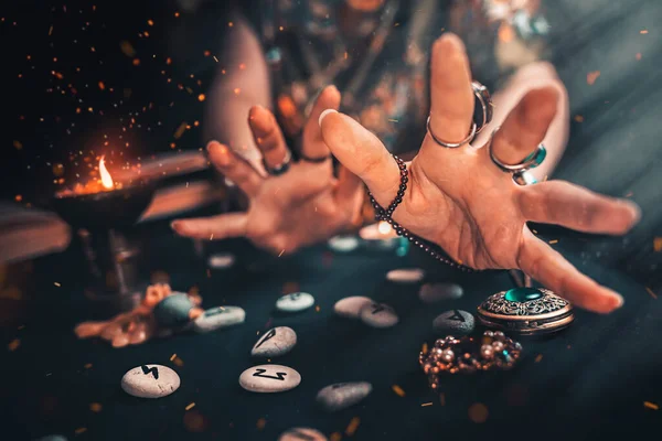 Astroloji ve ezoterizm. Cadı Runik Taşlardan okuyor ve yakınlarda bir mum var. Havada ateş kıvılcımları var. Karanlık arka plan. Avuçlar kapalı.
