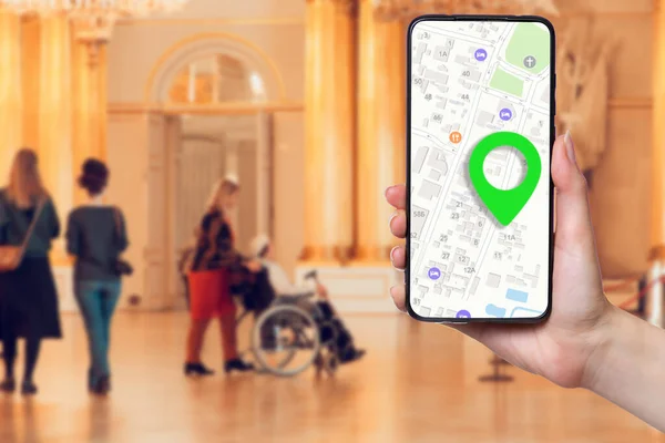 A females hand holds a smartphone with an online map on the screen. In the background, a group of people and a man in a wheelchair in a Museum, in a blur. Concept of online applications and navigation.