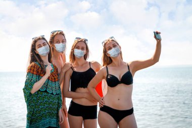 Virüs. Mayolu ve tıbbi maskeli bir grup güzel genç kadın denize karşı selfieler için poz veriyor. Virüs salgını sırasında deniz kenarında tatil kavramı.