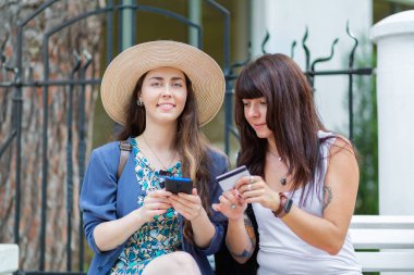 İki kadın sokakta oturuyor ve akıllı telefondan online bir mağazadan alışveriş yapıyorlar. Bir kadın elinde bir banka kartı tutuyor. İletişim ve modern teknoloji kavramı.