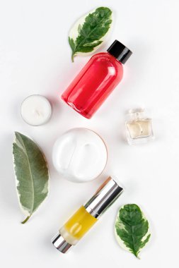 Kozmetik ve kişisel bakım. Yeşil yapraklar, duş jeli, parfüm, tüpteki yağ ve beyaz arka planda bir mum bileşimi serilmiştir. Dikey. Düz yatıyordu.
