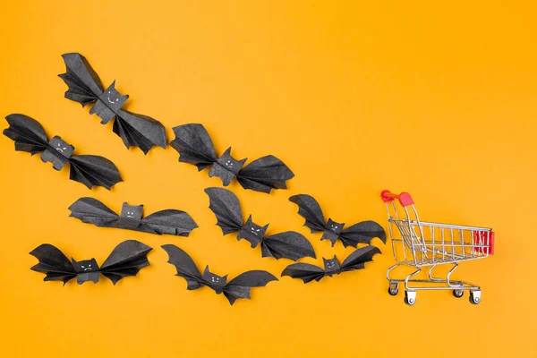 一群纸制的蝙蝠在购物车后面飞来飞去 橙色背景 复制空间 平躺在床上万圣节和假日购物的概念 — 图库照片