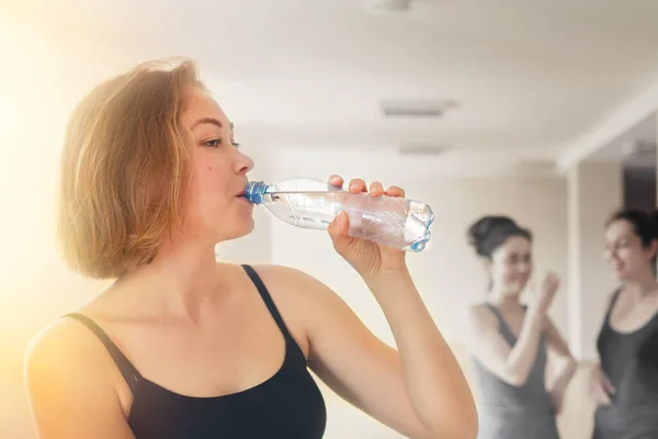 A young woman drinks water from a bottle. In the background are two women gossiping. The concept of sports training, socialization and communication.
