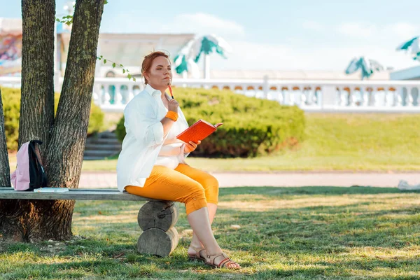 教育和商业 一个超重的女人坐在公园的长椅上 看上去很沉思 手里拿着记事本 — 图库照片