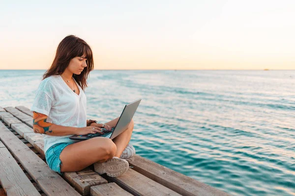 A young beautiful woman with tattoos is sitting on a pier by the sea and working at a laptop. Sea and sky in the background. Copy space. Concept of remote work and summer holidays.